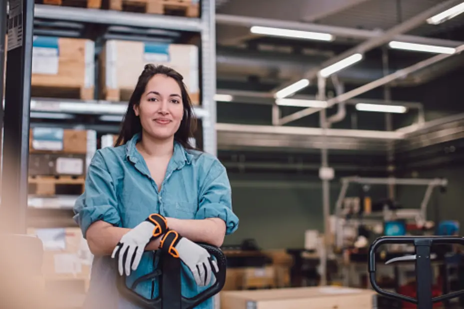 Жена с работно облекло и ръкавици позира усмихната пред склад, с кашони на заден план и палетна количка в близост.